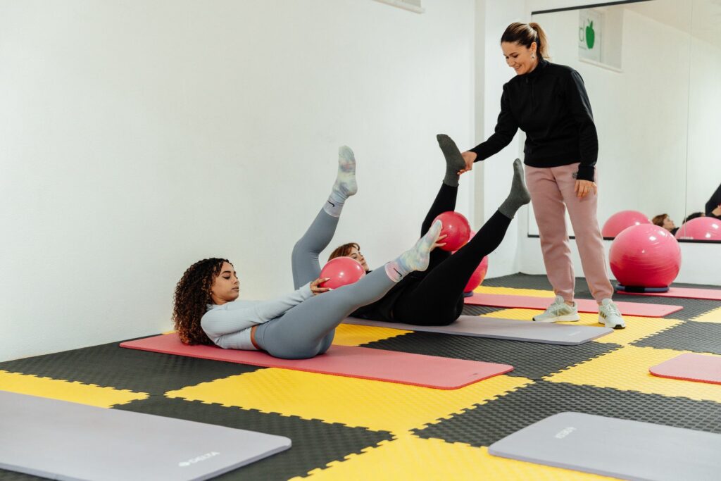 Cours de Pilates sur tapis : deux femmes allongées travaillent les abdominaux avec un petit ballon, guidées par une coach